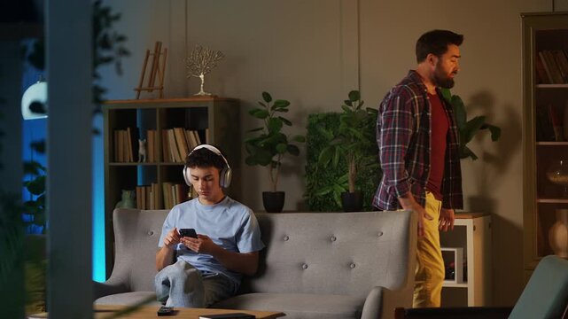 Grumpy dad shouting at rebellious teen son in headset using phone, conflict