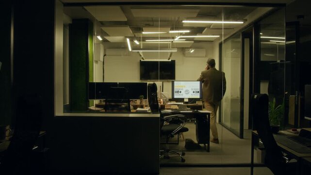 Wide shot through office window of mature Caucasian male manager working on financial report alone at midnight, answering phone call from wife, pacing around while arguing, work-life balance concept