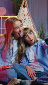 Attractive smiling carefree eldest and younger blond female relatives sitting on coverlet near decorative tent at home in the evening and photographing with amusing faces on smartphone