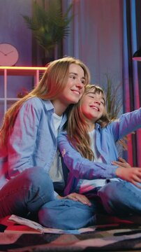 Good-looking active contented friendly blond native eldest and younger sisters relaxing on coverlets near decorative tent in nights light room interior and taking pictures theemselves on mobile