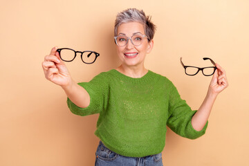 Mature woman smiles while holding glasses in green sweater against beige background for fashion...