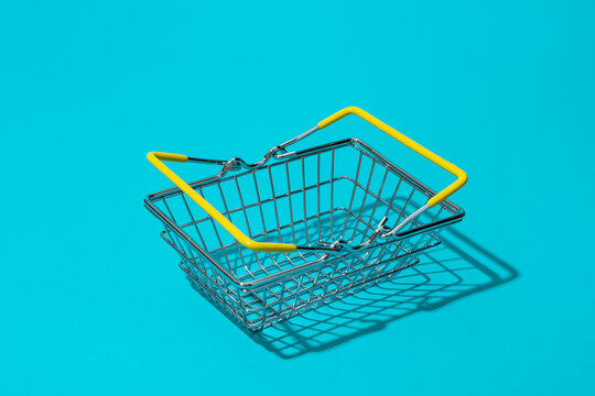 Empty metal shopping basket with yellow handles on bright turquoise blue background. Concepts of retail, shopping, consumer choice and sale season.