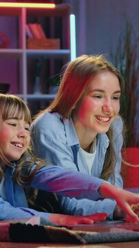 Leisure concept where adorable smiling carefree two girls of different ages lying on coverlets on the floor near decorative tent in playroom with night lights and talking during watching something on