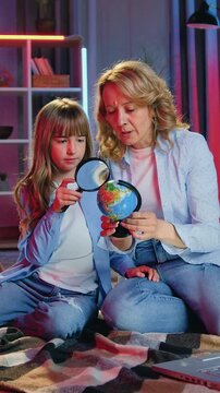 Home evening leisure concept where cute capable teen girl sitting on coverlets on the floor with lovely positive adult mom and examining mockup of earth through magnifying glass,front view
