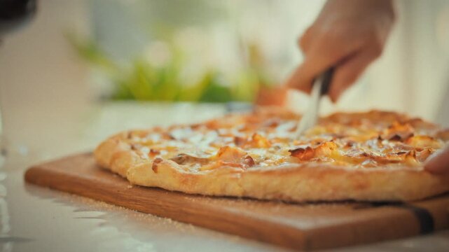 On bright day, housewife's skilled hands carefully cut homemade pizza on wooden board, using ordinary kitchen knife with precision ease, showcasing routine yet essential task in cozy kitchen setting