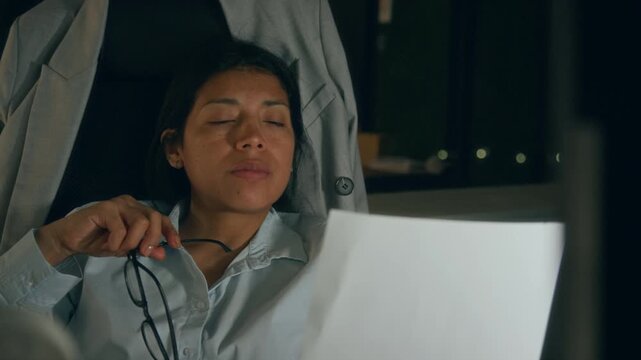 Medium close-up of frustrated, apathetic Hispanic female company CEO working alone in office at midnight, gnawing at glasses while analyzing document, then typing on computer