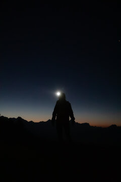 View of the silhouette of a hiker with a headlamp standing against the dark mountain peaks during twilight South Tyrol, Trentino-South Tyrol, Italy.