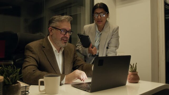 Medium shot of Caucasian male corporate executive with laptop and Hispanic marketing director with tablet discussing quarterly financial report while working to deadline late at night in dark office