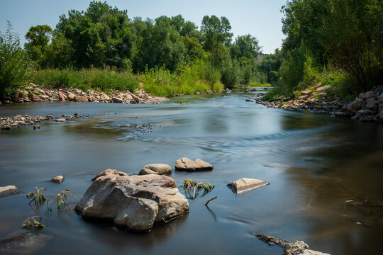Smooth surface on the Big Thompson River of Colorado.  Blue sky and green trees line the river. Trees in Colorado springtime with lush green foliage.  Glassy surface of long exposure.