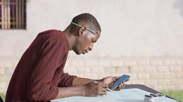 Black man planning painting using tablet outdoors, seated at table with paper and sketches, brick wall backdrop, sunlight on profile, pencil and pen ready, focused posture, creative layout session