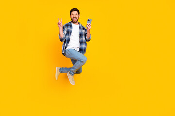 Obraz premium Happy man jumping with smartphone and peace sign on bright yellow background for social media, lifestyle and advertising concepts