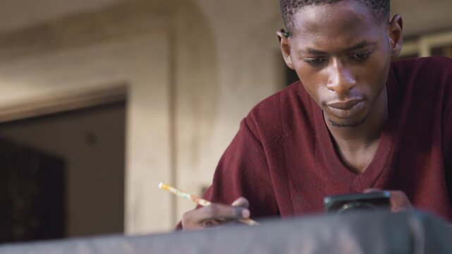 Black artist studying phone reference while sketching with pencil on work surface, leaning over table near doorway, focused expression, closeup portrait, natural light, thoughtful mood, creative