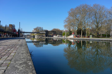 Fototapeta premium Paris, France - 03 15 2026: Ourcq Canal. Panoramic View of the canal with bridge, buildings and trees reflecting on the water from the quayside in sunny spring blue sky