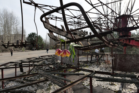View of the abandoned Pripyat amusement park with a rusty ride, two tulips hanging from a wooden seat, and people walking in the background in Pryp'yat', Kyiv Oblast, Ukraine.