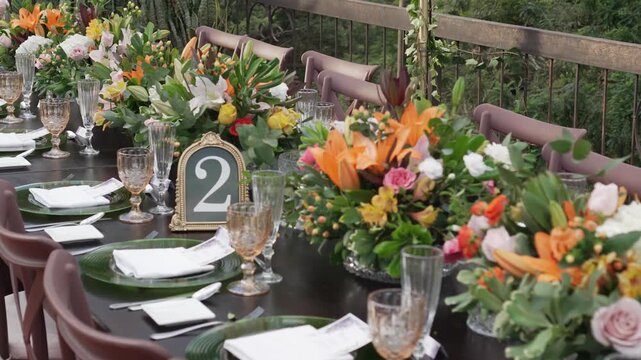 Decorated long table set for event with flower arrangements, table number two, green plates and crystal glasses, elegant outdoor celebration setup