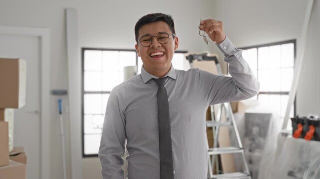 Man points finger to keyring while holding keys inside building with moving boxes and ladder; new home joy.