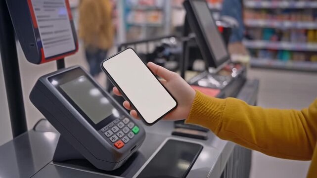 Close up of a person at a self service checkout in a supermarket using a mobile phone with a blank screen to make a contactless payment on a modern POS terminal for purchased goods