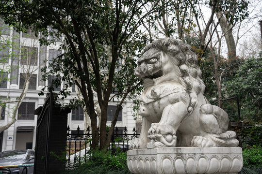 Stone lion sculpture in a courtyard with green trees and architectural background