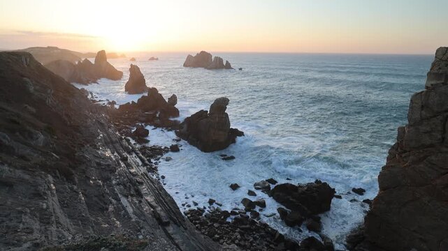 View of the dramatic sea stacks and jagged cliffs of Costa Quebrada with crashing waves under a golden sunset in Liencres, Cantabria, Spain.