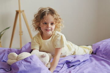 Fototapeta premium A young child with curly hair lies on a bed while holding a stuffed animal. The room is bright and cozy, with soft bedding and a relaxed atmosphere