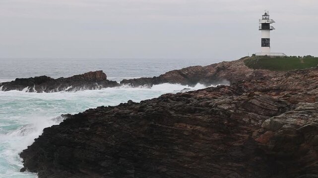 View of Faro Illa Pancha lighthouse standing on a rugged cliff with waves crashing against dark rocks under a cloudy sky in Isla Pancha, Lugo, Spain.