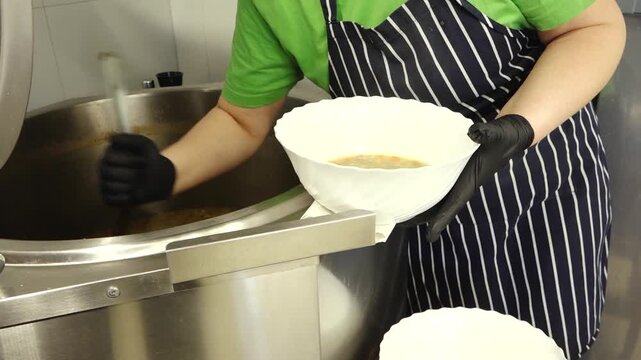 Close-up of a person ladling hot soup from a large metal pot into a white bowl, wearing gloves and apron in a kitchen or food service setting.