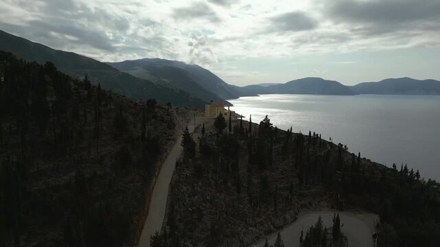 A winding road leads past a solitary yellow house on a tree-covered hill overlooking a calm sea and distant mountains under a cloudy sky.
