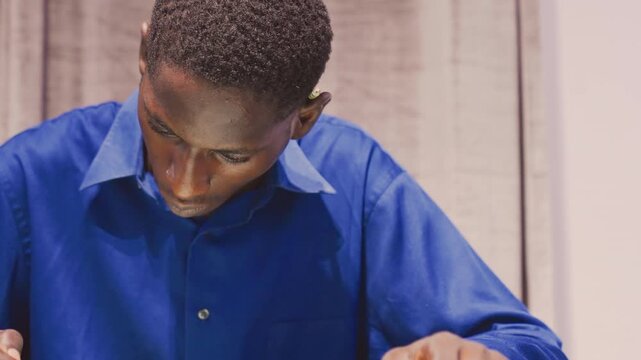 Young black man painting at desk blue shirt hands working with brush and pencil on paper and canvas, curtain backdrop, concentrated posture, steady strokes, reflective expression, intimate studio