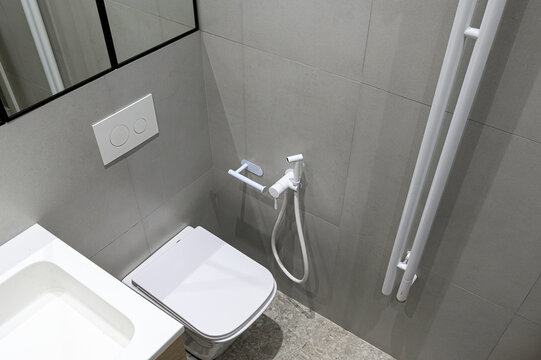 A modern bathroom with a wall-mounted toilet, bidet spray, heated towel rail, sink, grey tiled walls, minimalist design, flush panel above toilet, mirror partially visible.