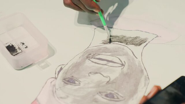 Overhead hand adding black ink to Black portrait on white paper, illustrator dips green tool into container, builds dark hair texture with stippling and dotted outline, smartphone reference nearby,