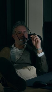 Full vertical shot of apathetic mature Caucasian male lawyer or manager sitting in dark office with feet resting on desk, staring at computer screen, while working on contract or business report