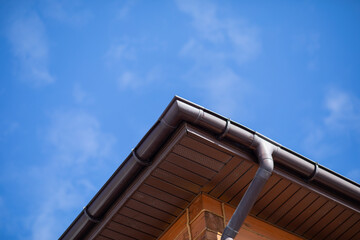 Brown gutter system on roof corner of wooden house against blue sky. Exterior drainage installation for rainwater management. Concept of home maintenance and residential construction. © Olesia Basova