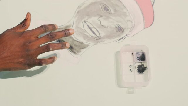 Black painting face being blended, Black conservator carefully smooths charcoal lines on portrait cutout using fingertips, palette and tray nearby, closeup of darkskinned hands retouching outline