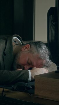 Vertical medium close-up of tired Caucasian businessman sleeping with head on desk in dark office at nighttime, while working on documents or financial report, suggesting burnout and rat race concepts