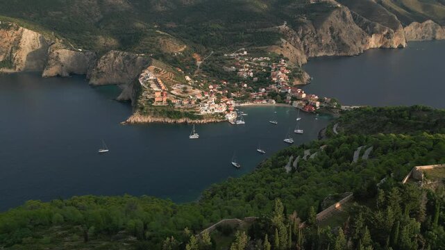 An aerial view of a picturesque village nestled between lush green hills and a calm, blue bay dotted with sailboats.