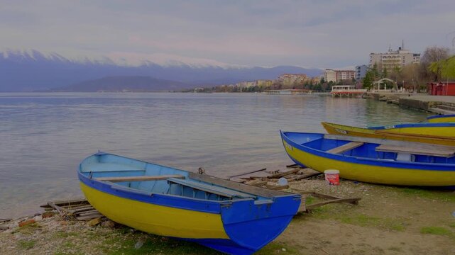 A panoramic and vibrant view of the Pogradec shoreline on Lake Ohrid, Albania. The photograph features traditional wooden fishing boats painted in bright blue and yellow resting on the rocky beach in 