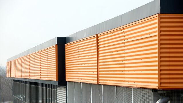 Architectural facade with orange cladding corrugated metal panels, large glass window of industrial or commercial building, contemporary architecture with vertical relief and material contrast.