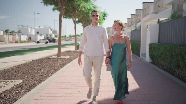 Young couple walking hand in hand on sunny street in modern residential area. Romantic outdoor lifestyle scene with love, relationship and summer city прогулка during vacation or daily life.