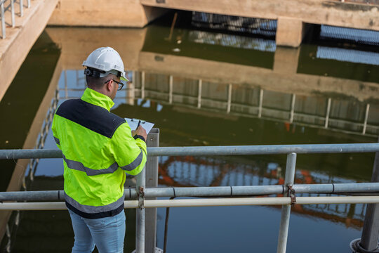 Professional Asian engineer inspecting water quality at a wastewater treatment plant. He is carefully recording maintenance data on a clipboard during an industrial site safety survey.