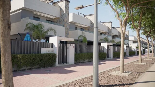 Peaceful residential street featuring a row of modern two-story townhouses with balconies, stone accents, and private gardens, lined by a sidewalk, trees, and streetlights