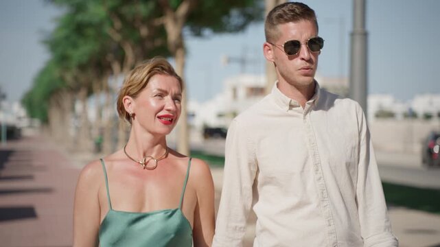 Mature woman and young man walking together on sunny city promenade. Romantic outdoor lifestyle scene with conversation, relationship and relaxed summer stroll in urban environment.