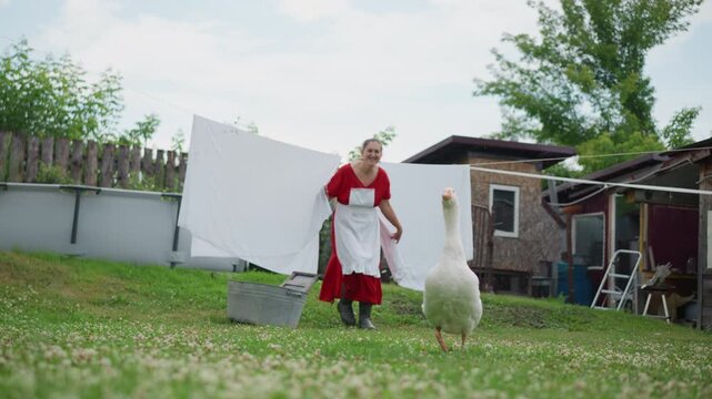 White woman walking beside clothesline goose, homestead caretaker guiding bird across lawn with linen flapping, cottage and trees framing scene, warm sunlight, slow gentle pace, human animal bond