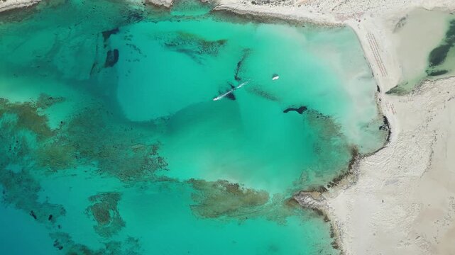 Cinematic top-down drone video of Balos Lagoon, where three seas meet. A lonely yacht and jet ski glide through vibrant shades of turquoise, azure, and deep indigo. High quality 4k footage