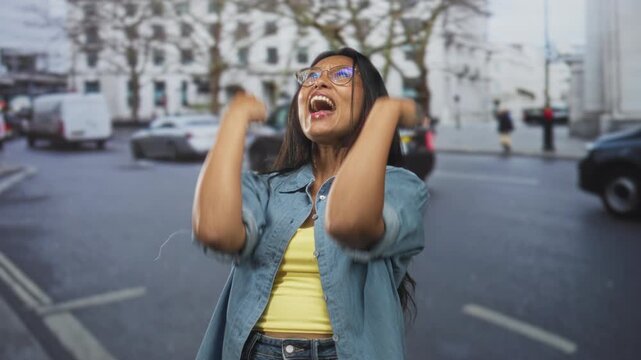 Woman clenches fists to chest and laughs with closed eyes beside moving cars on a city street, wearing denim shirt, yellow top and glasses; joy celebration.