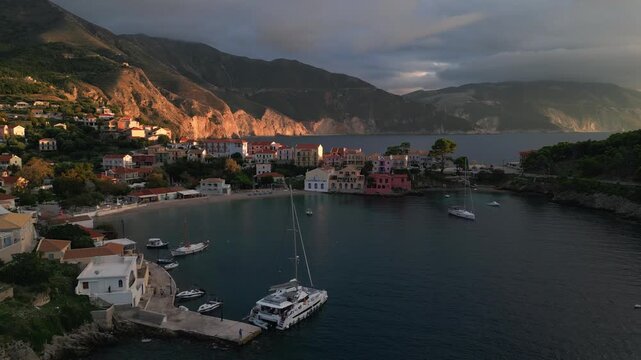 A picturesque Greek coastal village nestled against a mountain under a dramatic, cloudy sky, with sailboats dotting the calm sea.