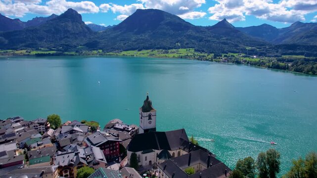 Aerial 4K View of St. Wolfgang Village and White Church. Cinematic Drone Flyover towards Turquoise Lake Wolfgangsee and Alpine Mountains. Scenic Waterfront with Small Boats. Salzkammergut Austria