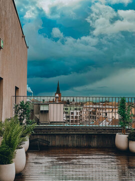A rain-drenched rooftop terrace overlooks a European city, a church spire and a dramatic reflection of a cloudy sky.