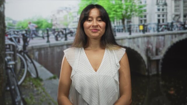 Hispanic young woman smiling, lip piercing visible, wearing white top by a canal bridge on a street in amsterdam, relaxed posture and soft gaze; urban quiet contentment.