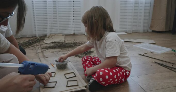 Young woman teaching two children how to make art from natural materials, creating small frames by gluing together wooden twigs while sitting together on the floor in a creative workshop.