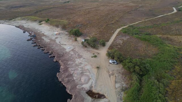 Aerial view of Paralia Skidi beach on Limnos Island, Greece, featuring a parked campervan beside a remote coastline. Clear Mediterranean water and rugged terrain highlight peaceful wild camping
